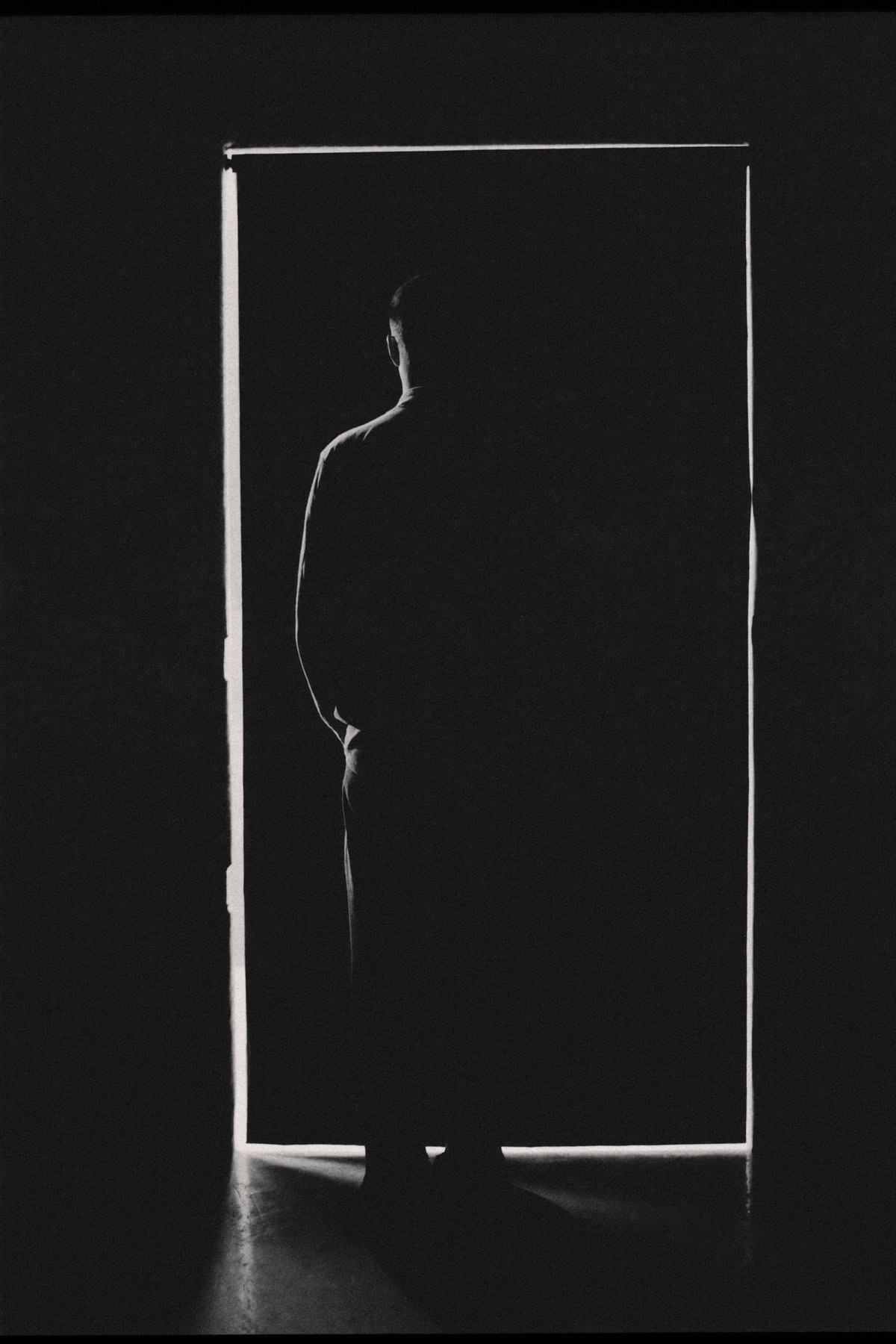 Anonymous figure seen from behind in a dark doorway, rim light tracing the silhouette